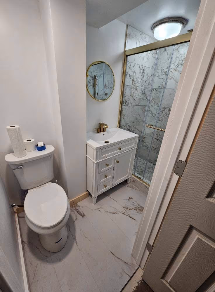 Finished compact bathroom with a glass shower, white vanity, gold fixtures, and marble-look floor tile