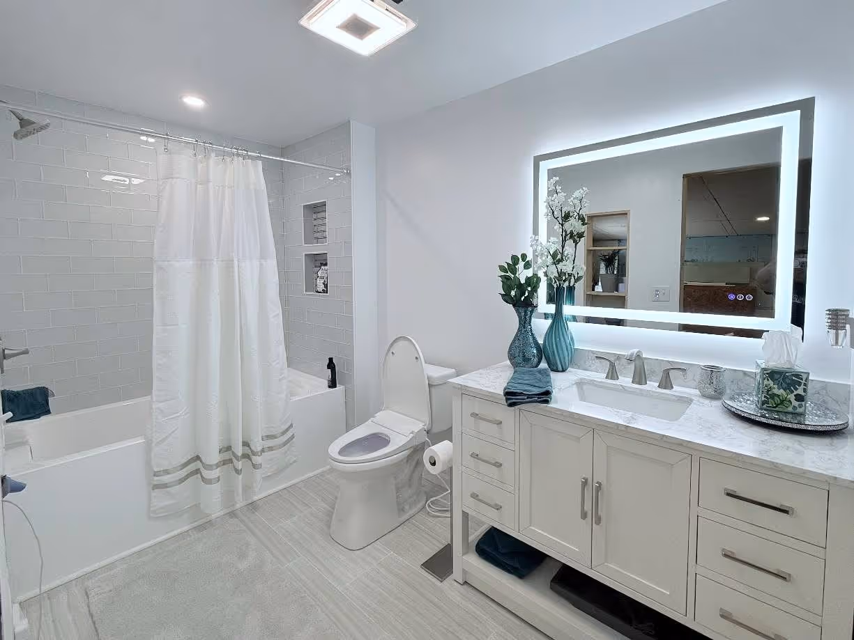 Finished bright bathroom with a white double vanity, illuminated mirror, bathtub, and large-format floor tile