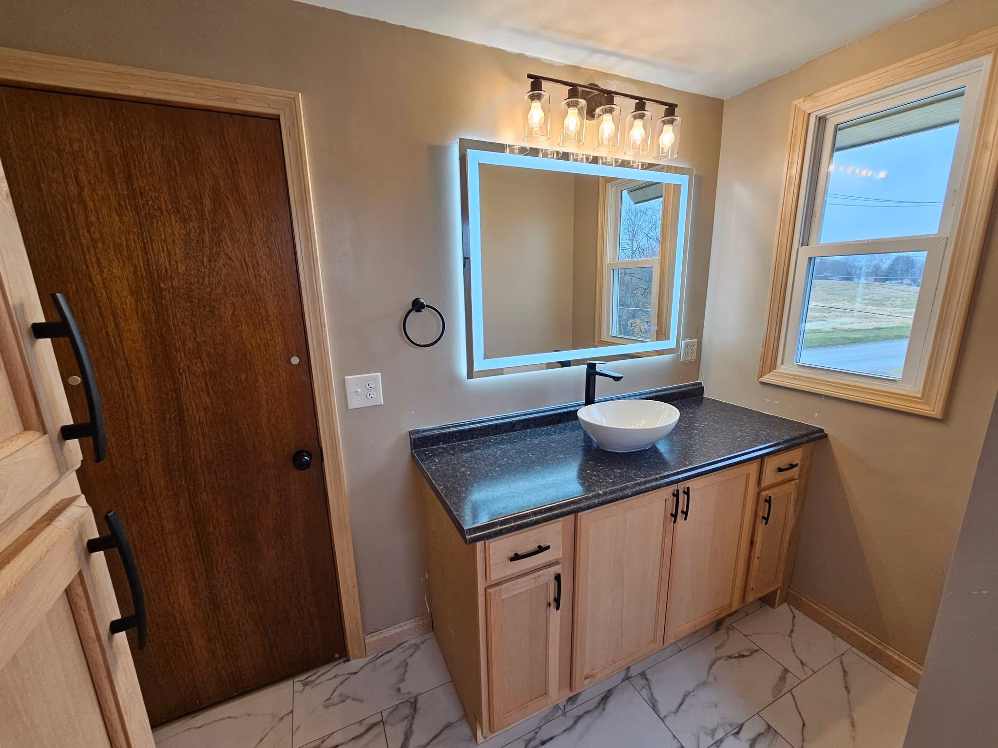 Finished bathroom with a natural wood vanity, vessel sink, illuminated mirror, and marble-look floor tile